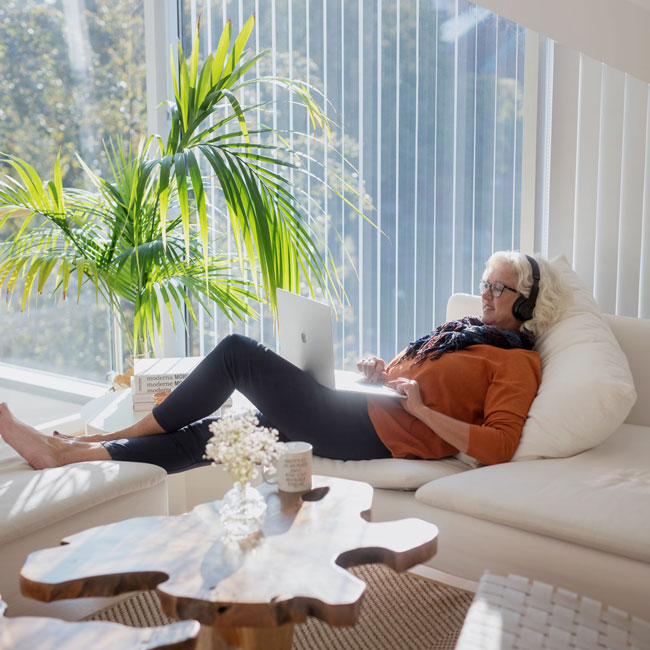 Eldre dame med som sitter på en hvit sofa med en laptop i fangen, mens hun hører på musikk med hodetelefoner.